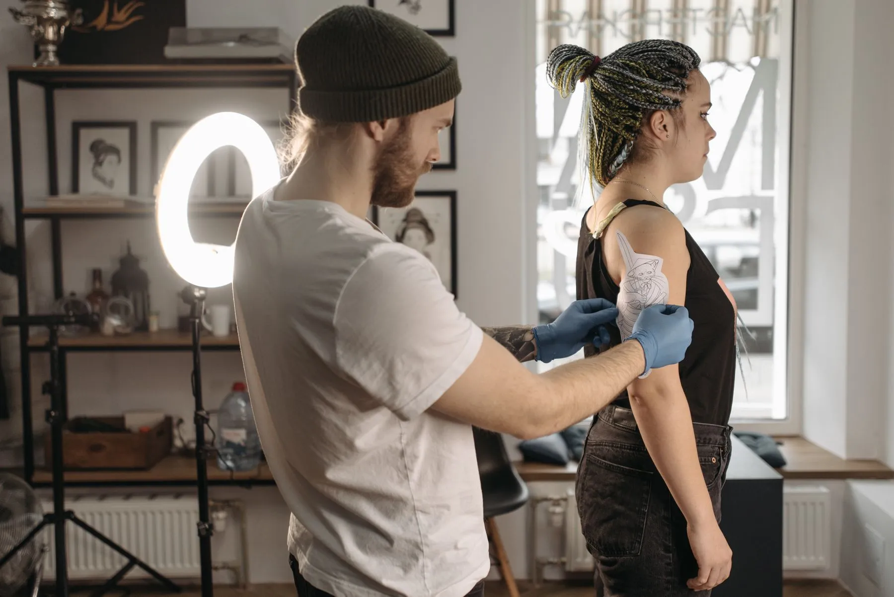 An artist checking stencil placement on a client's upper arm in a tattoo studio